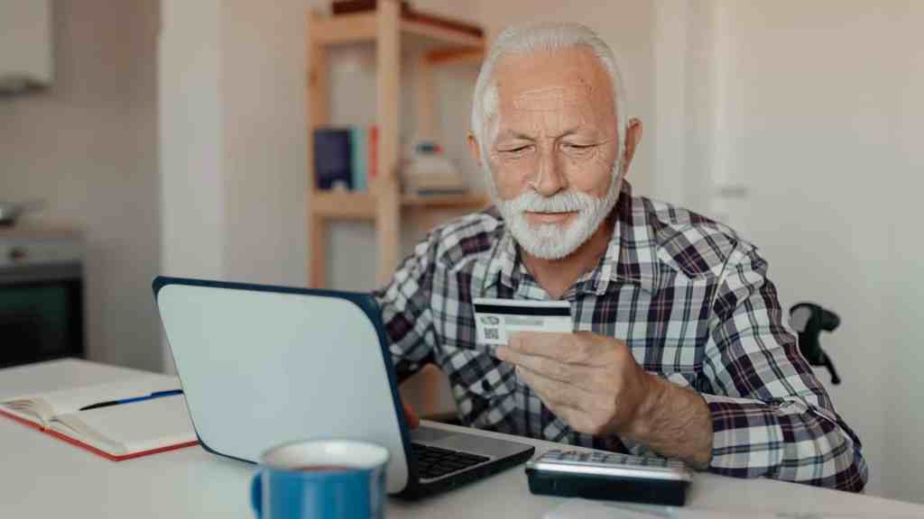 Ein älterer Herr sitzt mit dem Smartphone in der Hand vor einem Laptop.