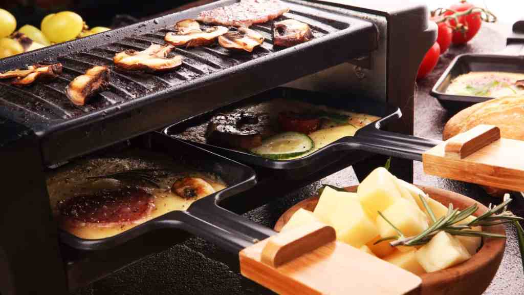 Nahaufnahme eines Raclette-Grills, auf der Grillplatte liegen Pilze, die Pfännchen sind befüllt und mit Käse überdeckt