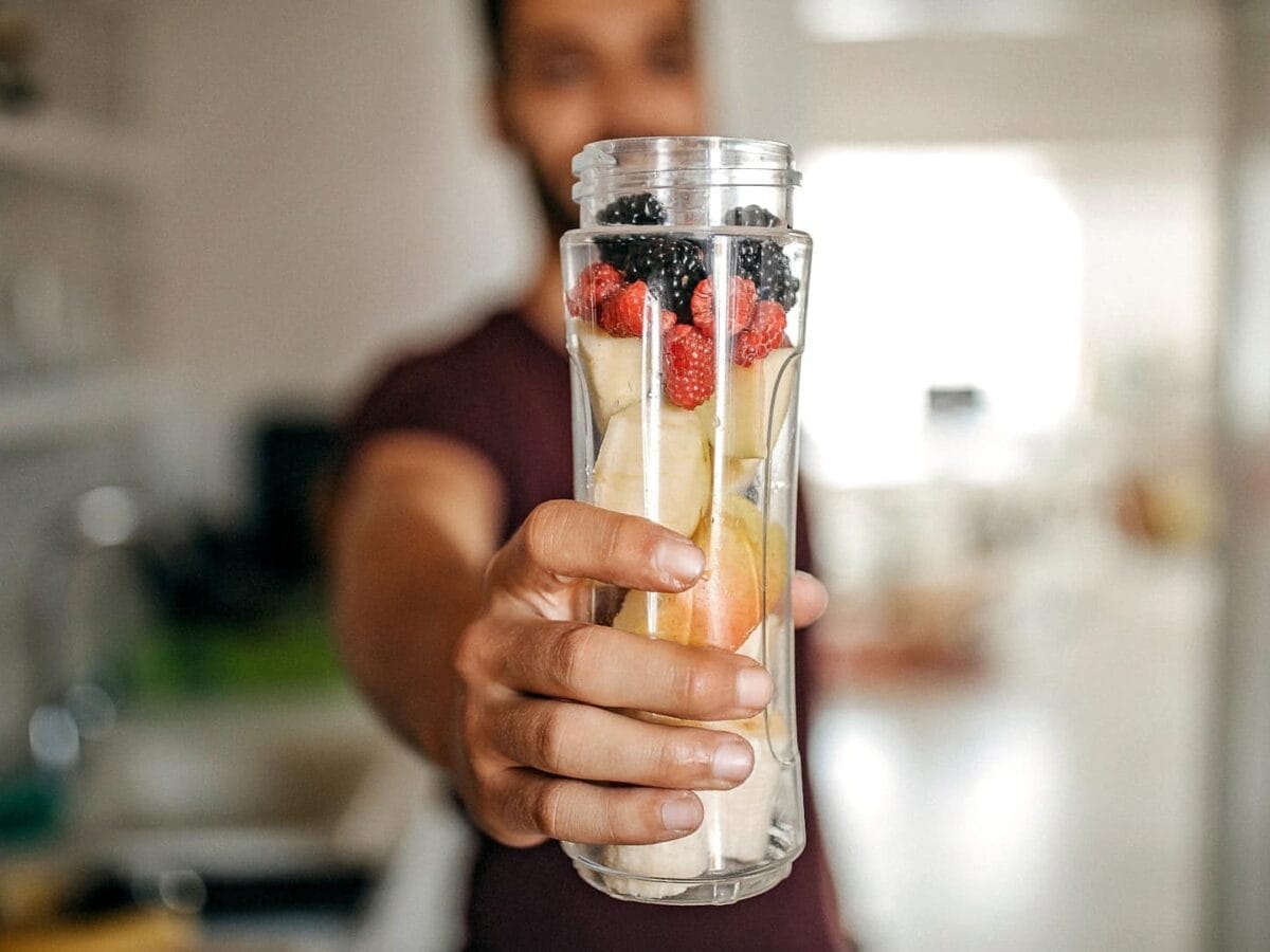 Mann hält einen Smoothiemixer voll Früchten in die Kamera.