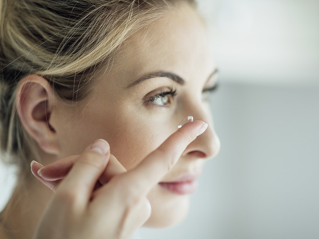 Blonde Frau hält mit Finger eine Kontaktlinse ans Auge, Profil schräg von der Seite