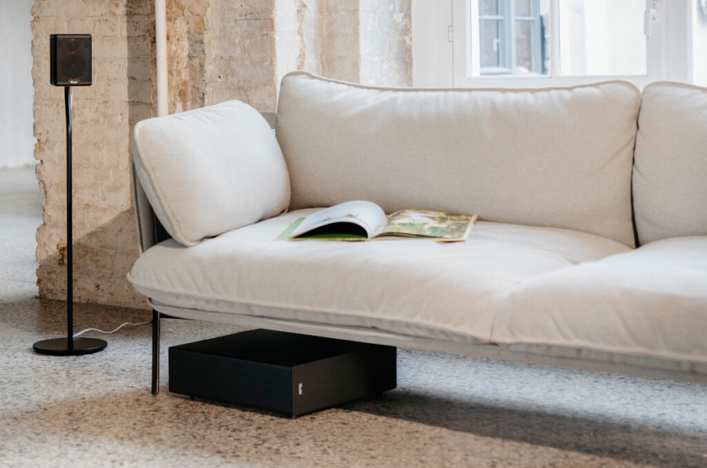 Weißes Sofa mit liegendem schwarzen Subwoofer drunter, daneben ein Lautsprecher auf Ständer