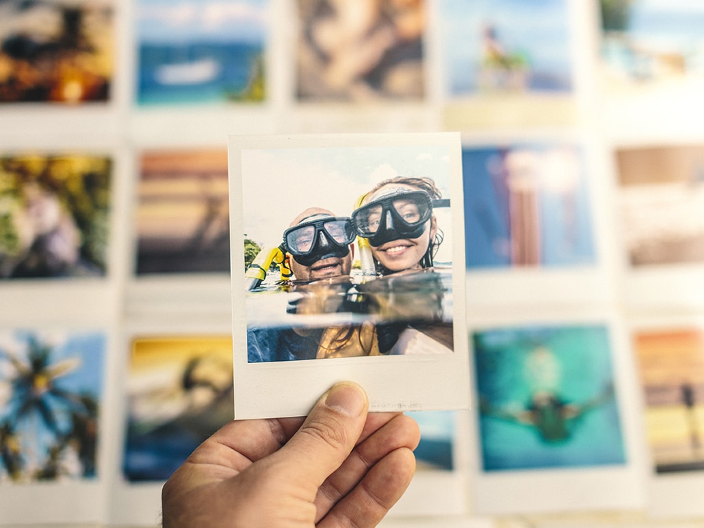Hand hält ausgedrucktes Urlaubsfotos von zwei Kinderköpfen mit Taucherbrillen im Wasser, im Hintergrund liegen viele weitere in Reihen angeordneten Fotos