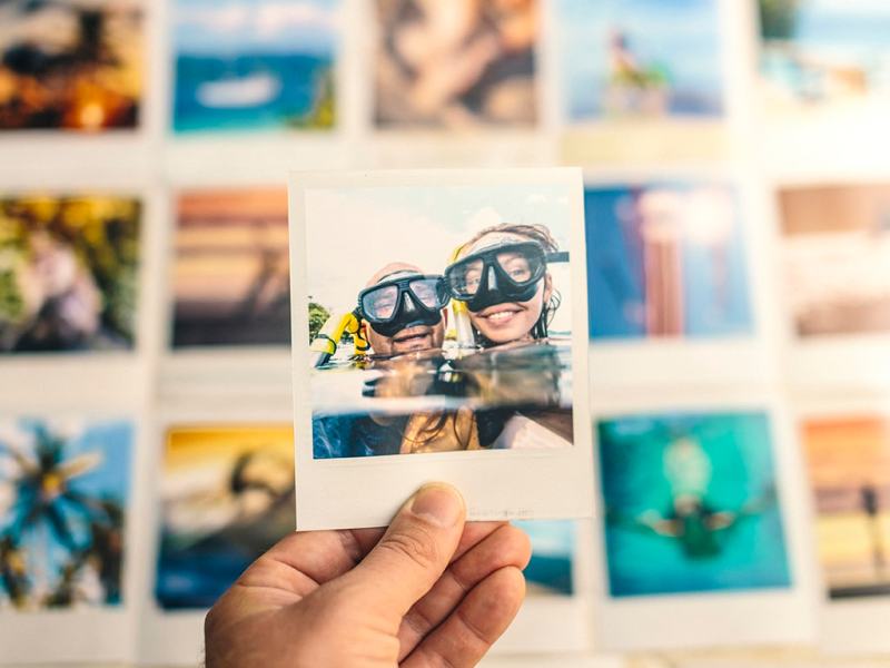Hand hält ausgedrucktes Urlaubsfotos von zwei Kinderköpfen mit Taucherbrillen im Wasser, im Hintergrund liegen viele weitere in Reihen angeordneten Fotos