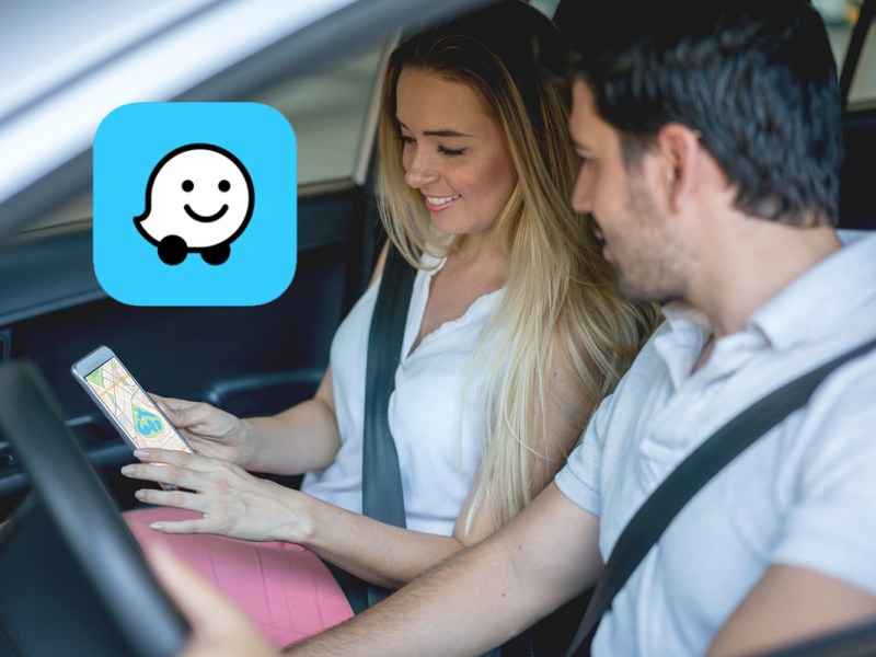 Junges Paar sitzt lächelnd im Auto und schaut auf Smartphone, das die Frau hält
