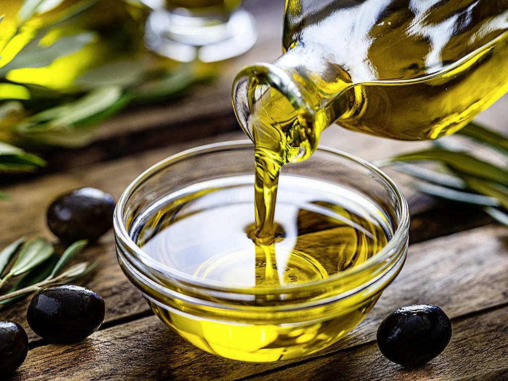 Olivenöl wird aus Glasflasche in Glasschüssel gekippt, steht auf Holztisch mit Olivenzweige drauf