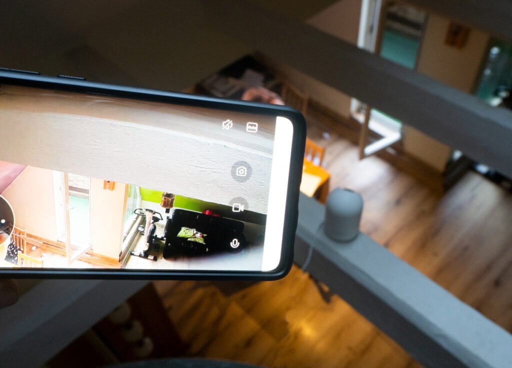 Smartphone ragt von links ins Bild und zeigt Aufnahme der Überwachungskamera, die dadrunter auf einem Balken platziert ist