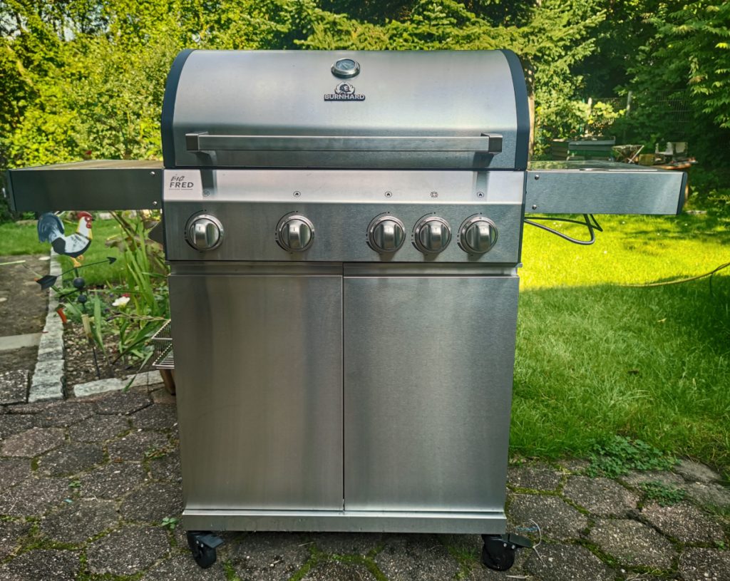 Silberner Gasgrill von vorne auf Terrasse umgeben von grünem Rasen und Pflanzen