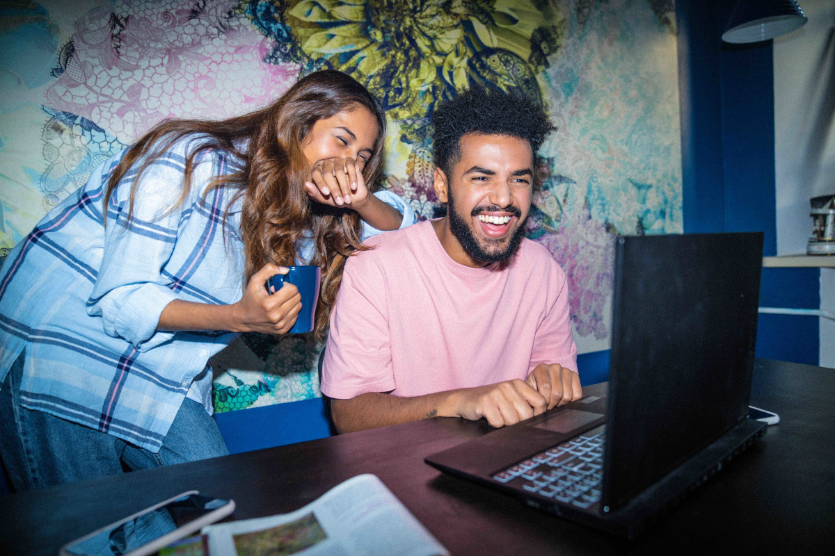 Junge Frau und junger Mann sitzen lachend vor Laptop
