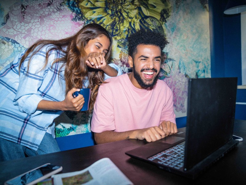 Junge Frau und junger Mann sitzen lachend vor Laptop