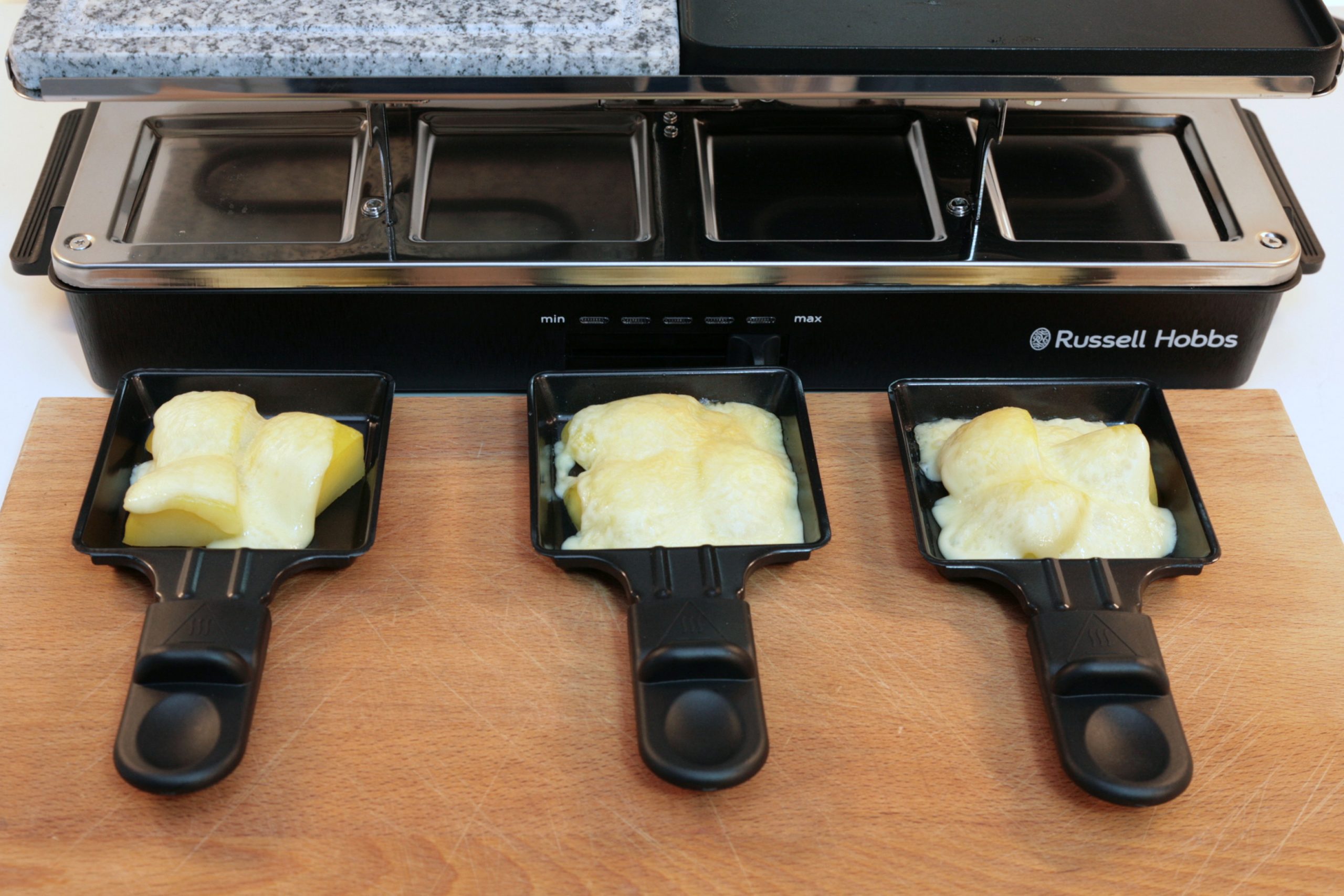 Drei Raclette Pfannen mit geschmolzenem Käse vor Grill