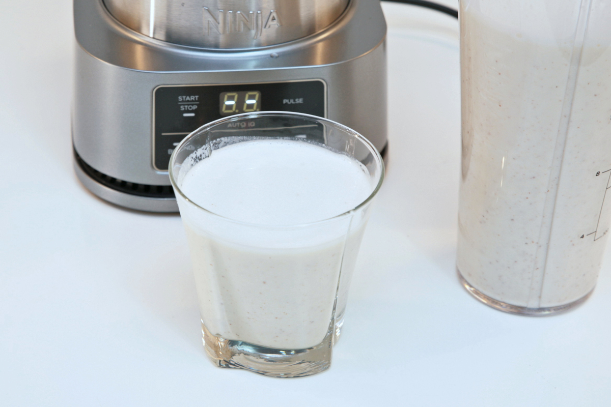 Kleiner Becher mit weißem Smoothie vor silbernem Standmixer