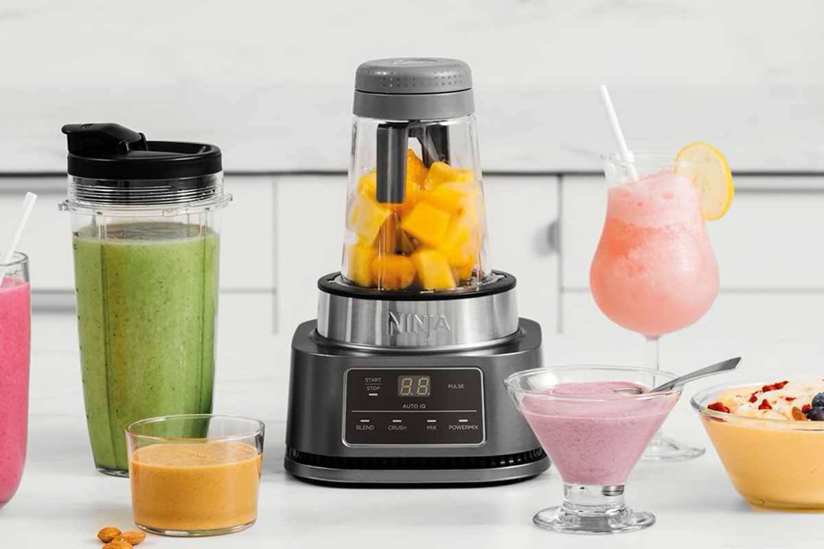 Grauer Standmixer in der Mitte mit gelben Stücken Obst, daneben verschiedene Becher, Gläser und schüsseln mit unterschiedlich farbigen Smoothies, Saucen und Getränke