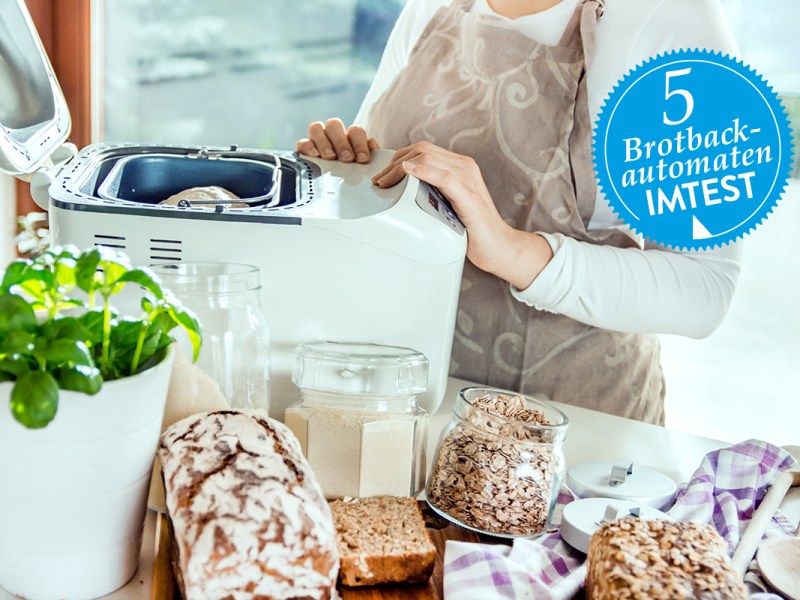 Frau ab Hals abwÃ¤rts im Bild hÃ¤lt HÃ¤nde an weiÃŸem geÃ¶ffneten Brotbackautomaten, der auf Tisch mit Brot und Zutaten steht