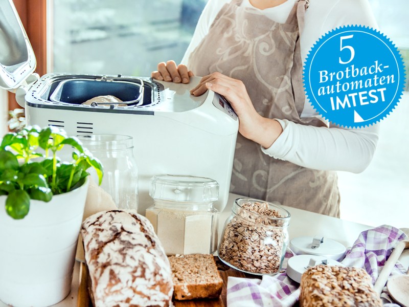 Frau ab Hals abwärts im Bild hält Hände an weißem geöffneten Brotbackautomaten, der auf Tisch mit Brot und Zutaten steht