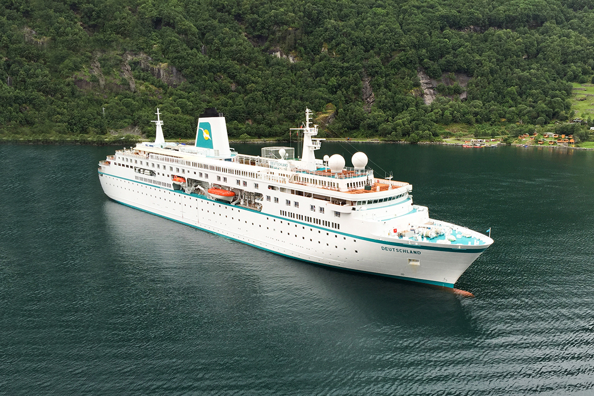 Blick schräg von oben auf Wasser mit weißem Kreuzfahrtschiff vor grüner Küste