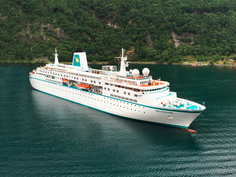 Blick schräg von oben auf Wasser mit weißem Kreuzfahrtschiff vor grüner Küste