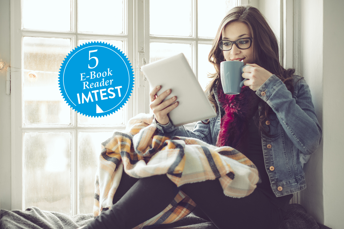 Frau mit Brille sitzt mit eBook-reader und Tasse in den Händen warm angezogen auf der Fensterbank