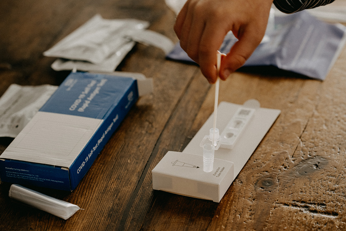 Ausgepackter Corona-Schnelltest auf Holztisch; Hand hält Teststäbchen in aufgestelltes Teströhrchen