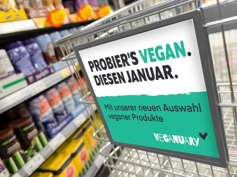 Ecke Einkaufswagen mit grün weißem schild vorne dran von Veganuary, im Hintergrund ein Regal im Supermarkt