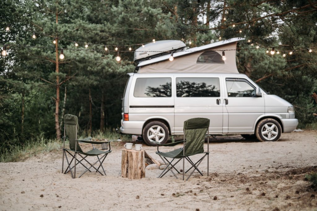 Weißer Camping-Van mit ausgeklapptem Zeltdach auf Sandboden vor grünen Bäumen. Campingstühle davor