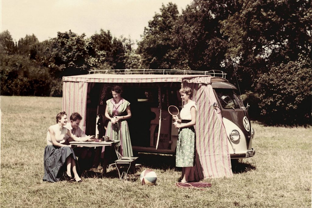 Menschen vor einem VW-Oldtimer (VW Bus T1 mit Westfalia-Campingausbau) stehend auf einer Wiese.