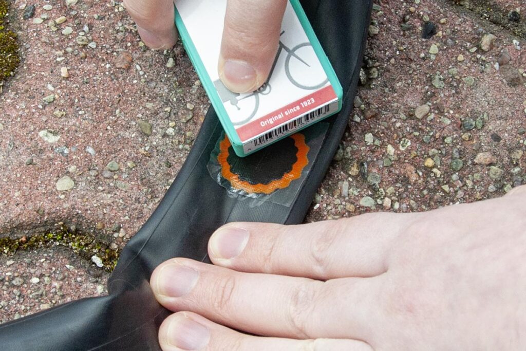 Hand drückt Reifenschlauch auf Boden während andere Hand mit türkisem Teil Flicken aufbringt