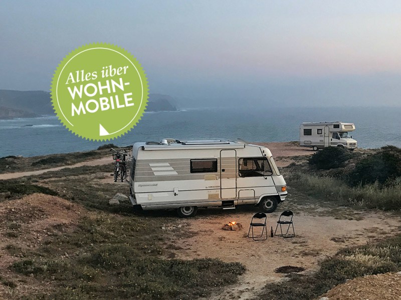 Zwei Wohnmobile stehen im Abendlicht auf einer Landzunge über dem Meer. Vor einem Wohnmobil brent ein kleines Lagerfeuer