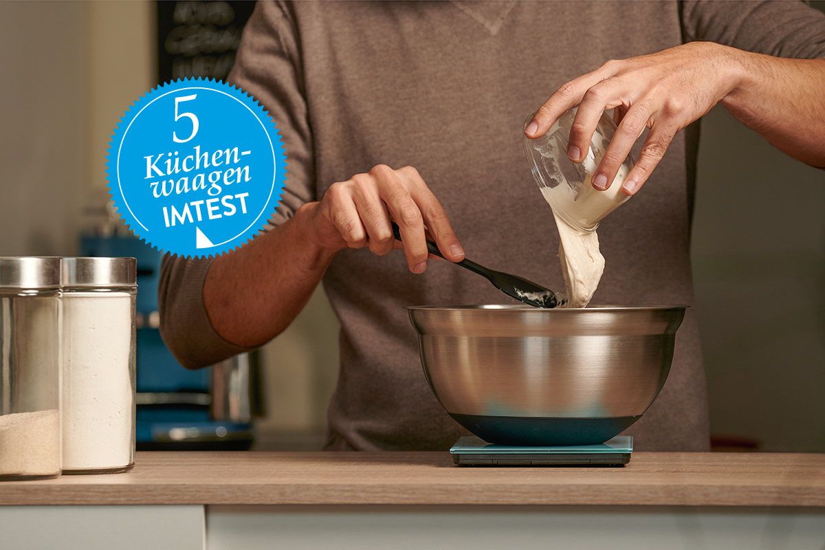 Hände von Mann schütten Mehl in Schüssel, die auf Küchenwaage auf Theke steht; blauer Sticker mit Küchenwaagen-Test