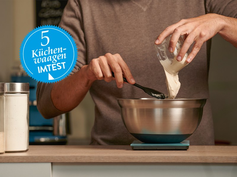 Hände von Mann schütten Mehl in Schüssel, die auf Küchenwaage auf Theke steht; blauer Sticker mit Küchenwaagen-Test