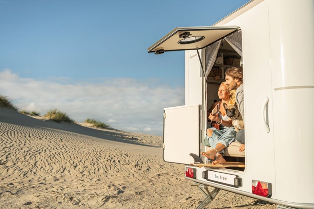 Man und Frau sitzen in einem Wohnwagen, der am Strand steht und schauen bei geöffneter Klappe in die Sonne.
