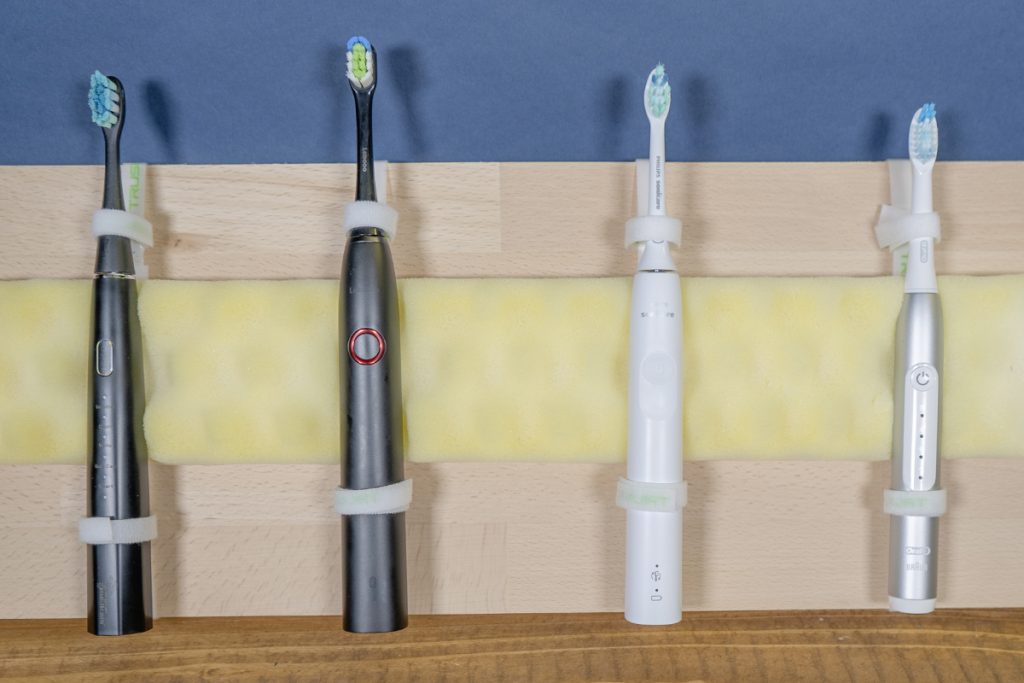 Vier verschiedene elektrische Zahnbürsten mit Kabelbindern senkrecht an Holz befestigt