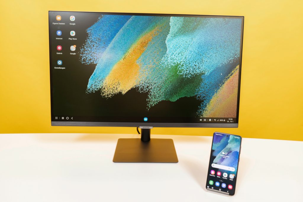 Home-Office-Monitor mit buntem Hintergrund auf Tisch, davor aufgestelltes Smartphone mit Startbildschirm