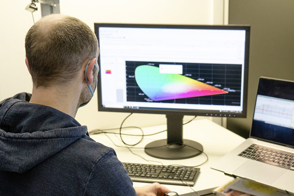 Mann schräg von hinten sitzt vor Computer, der ein buntes Diagramm zeigt während er Messgerät über Smartphone hält