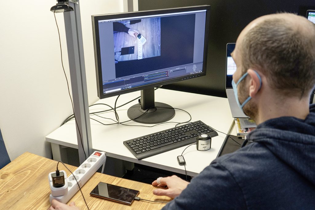 Mann von schräg hinten vor Computerbildschirm mit angeschlossenem Smartphone