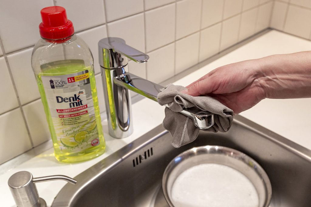 Hand mit grauem Lappen putzt Wasserhahn von Spülbecken, daneben flasche mit gelben Reiniger