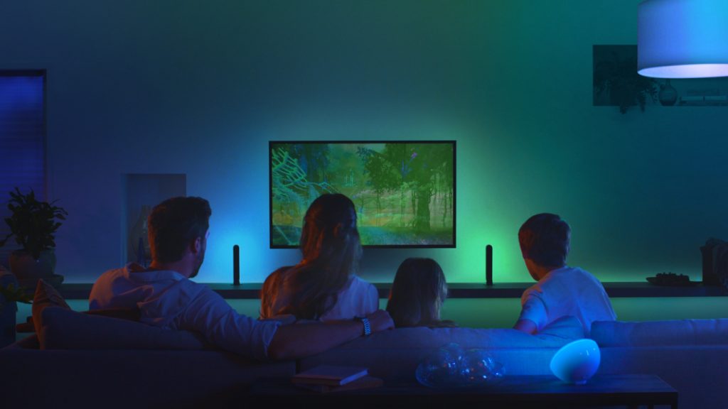 Familie von hinten sitzt auf Sofa in dunkelblau beleuchtetem Zimmer vor dem Fernseher, der ein grünes Bild zeigt