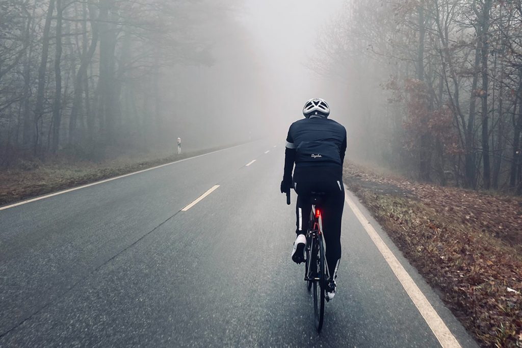 Rennradfahrer unterwegs bei Regen auf einer Strasse, die durch einen Wald führt.