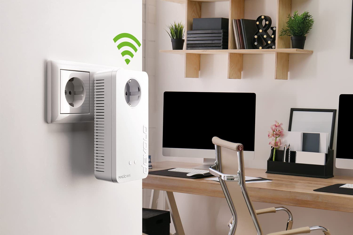 Weißer Steckdosen Adapter an Wand im Vordergrund mit grünem Wifi-Symbol, im Hintergrund Schreibtisch mit Monitor und Pflanzen