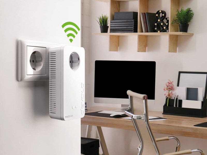 Weißer Steckdosen Adapter an Wand im Vordergrund mit grünem Wifi-Symbol, im Hintergrund Schreibtisch mit Monitor und Pflanzen