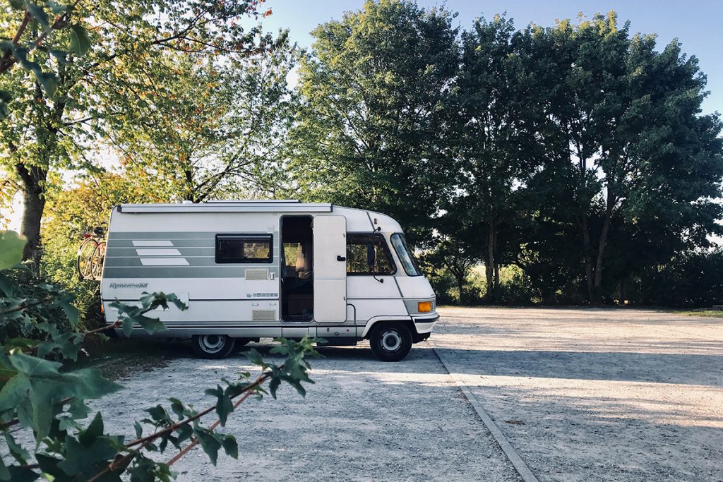 Wohnmobil steht mit offener Tür zum Wohnraum auf einem Parkplatz unter Bäumen.