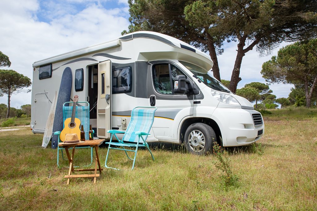 Wohnmobil mit geöffneter Tür auf einem Stellplatz vor Bäumen. Tisch, Stühle, Sufbrett und Gitarre stehen außerhalb des Campers auf der Wiese.