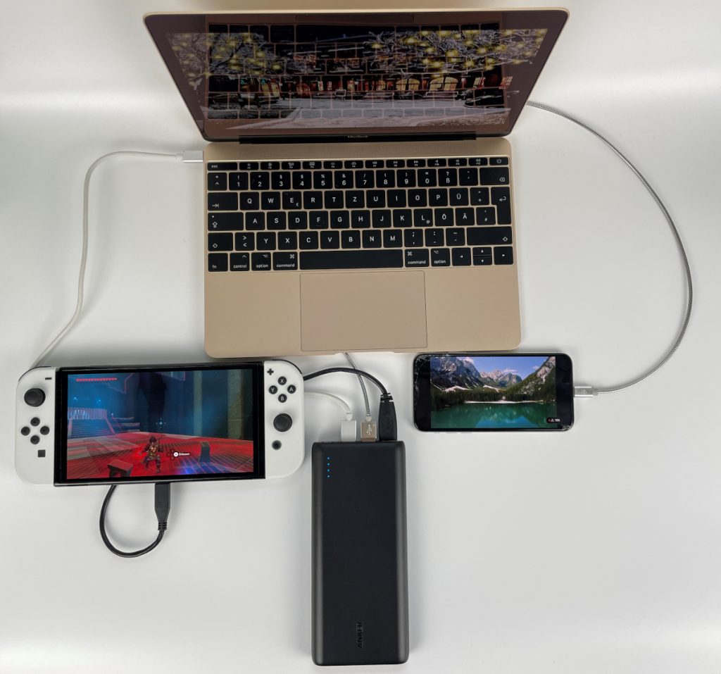 Powerbank von oben angeschlossen an weißer Nintendo Switch, goldenem Notebook und schwarzem Smartophone