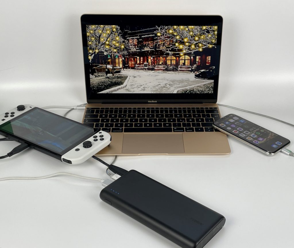 Vorne schwarze Powerbank, links weiße Switch, in der Mittel goldenes Macbook und rechts ein Smartphone auf weißem Tisch. Alles verkabelt