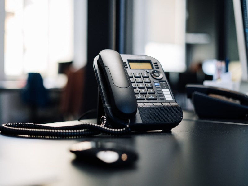 Schwarzes Festnetztelefon auf Bürotisch von Kundenservice, im Hintergrund andere Telefone und Computer