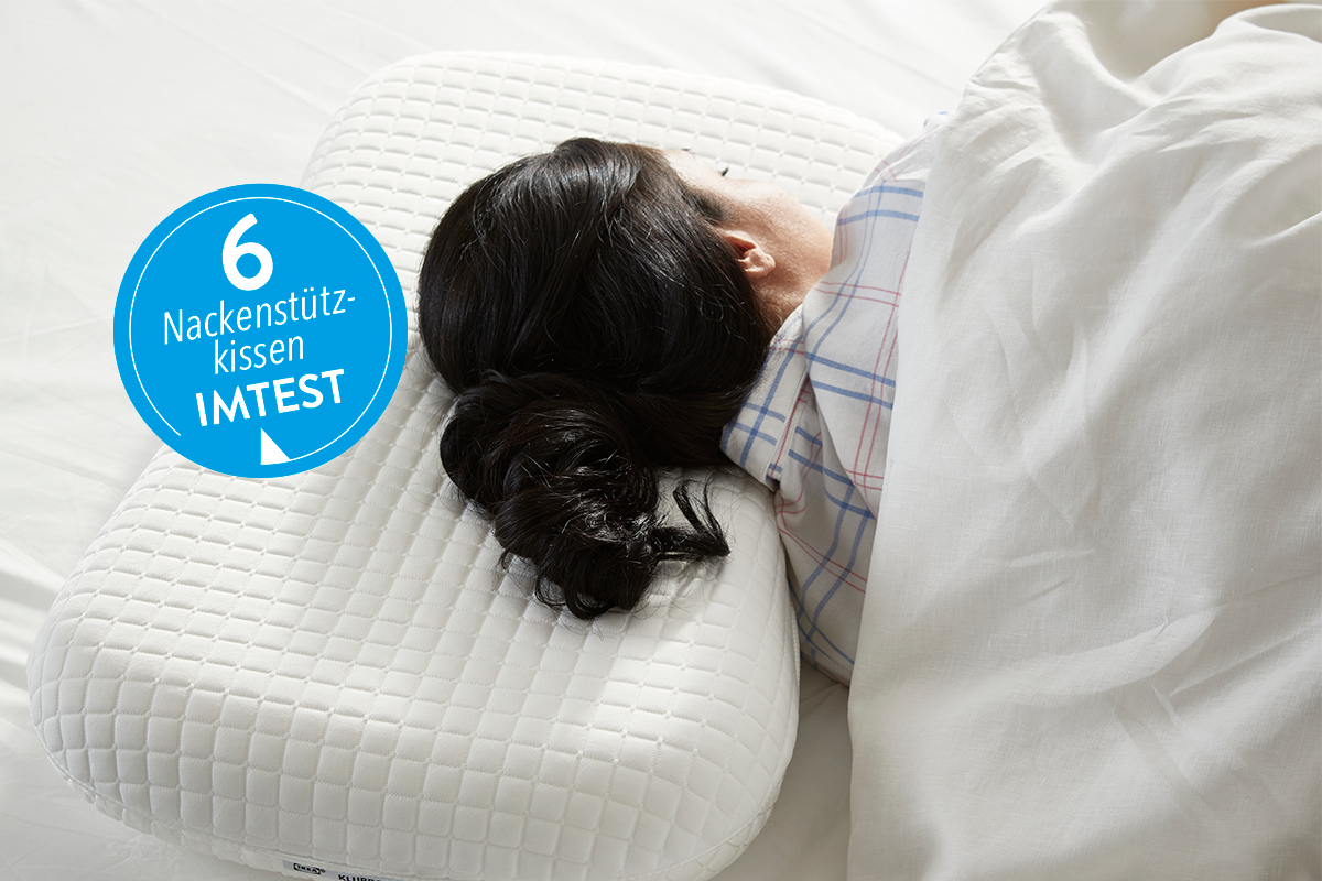 Person mit langen schwarzen Haaren schläft unter weißer Decke auf Nackenstützkissen; blauer Sticker mit "6 Nackenstützkissen IMTEST"