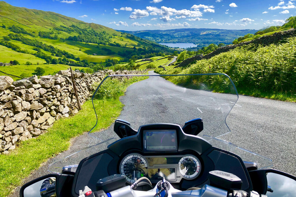Straßenverlauf aus der Sicht eines Motorradfahrers – in der Ferne sind Berge uns ein See zu sehen.