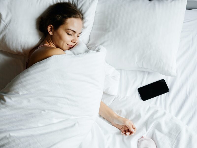 Frau mit weißer Decke zugedeckt im Bett lächelt mit geschlossenen Augen, neben ihr ein Smartphpne
