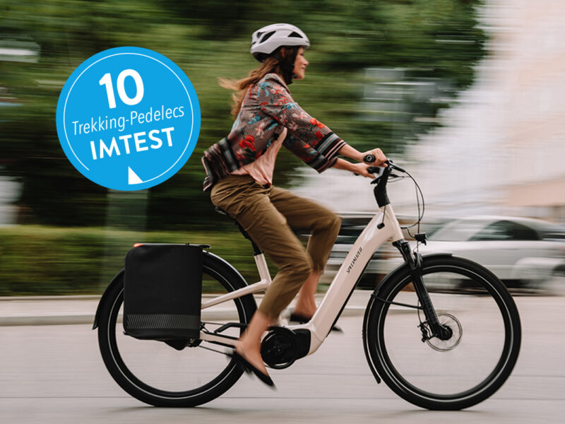 Frau fährt auf weißem Trekking-E-Bike in der Stadt;  links ein blauer Aufkleber mit 