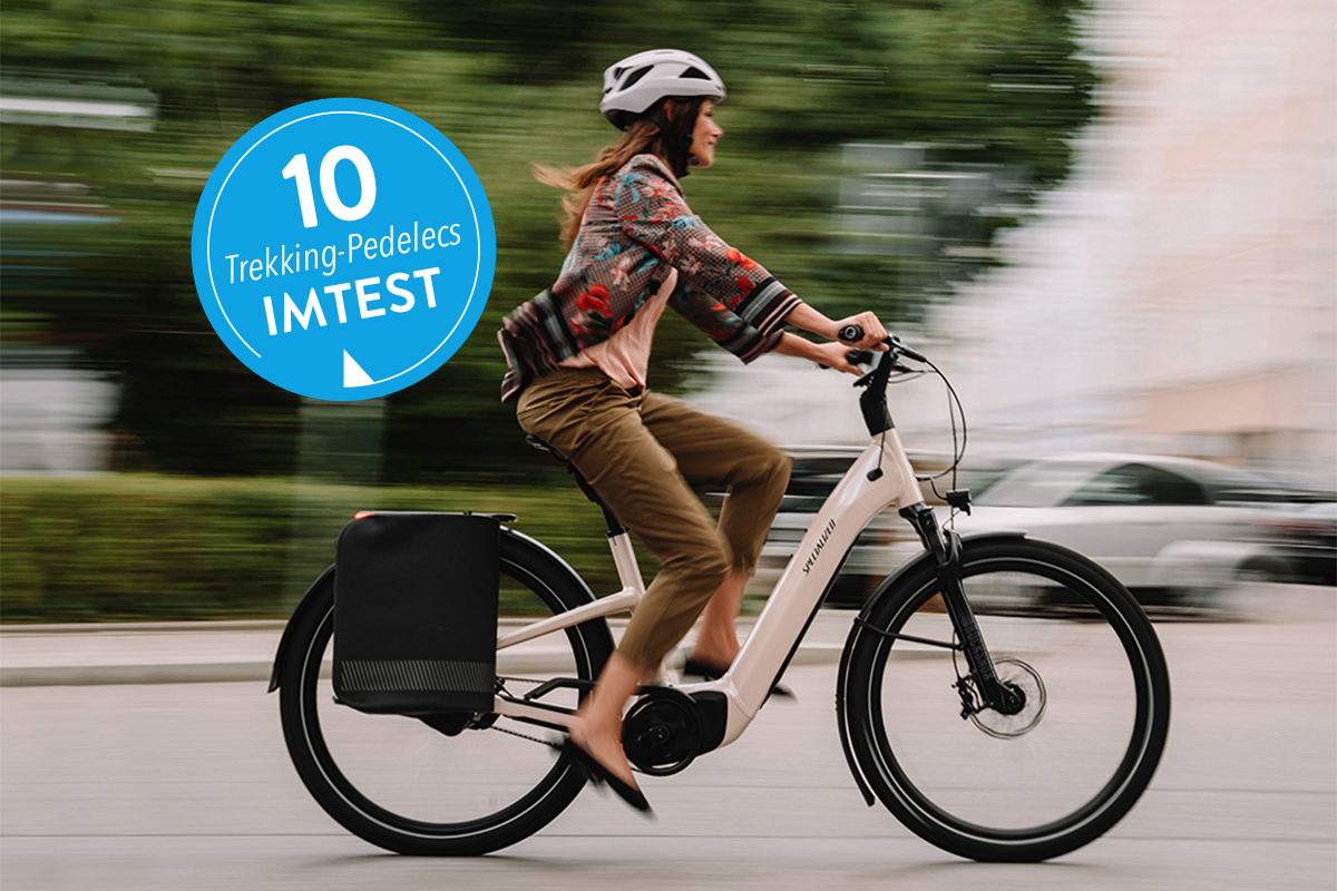 Frau fährt auf weißem Trekking-E-Bike in der Stadt; links ein blauer Sticker mit "10 Trekking-Pedelecs IMTEST"
