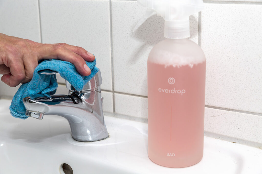 Hand mit blauem Tuch putzt Wasserhahn in Bad, daneben Glas-Sprühflasche mit orangener Flüssigkeit
